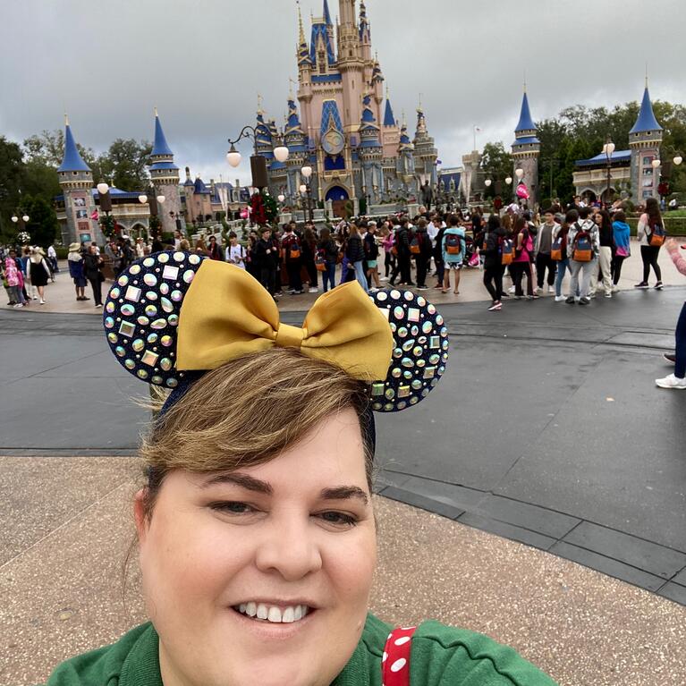 Heather Morales smiles while wearing Minnie Mouse ears in front of the Disneyland Castle on a cloudy day