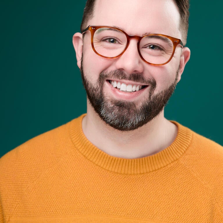 Joshua Carter smiles while wearing an orange sweater and brown glasses while standing agains a hunter green background
