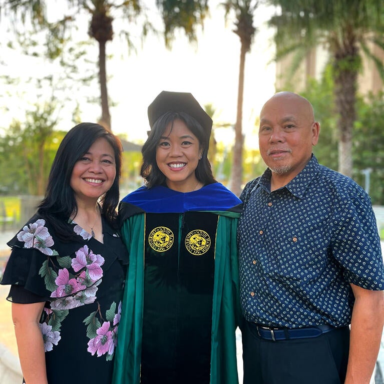 Advancement Ally, Christina Magalona, poses in her commencement regalia and is flanked by her parents on either side. There are palm trees in the background.