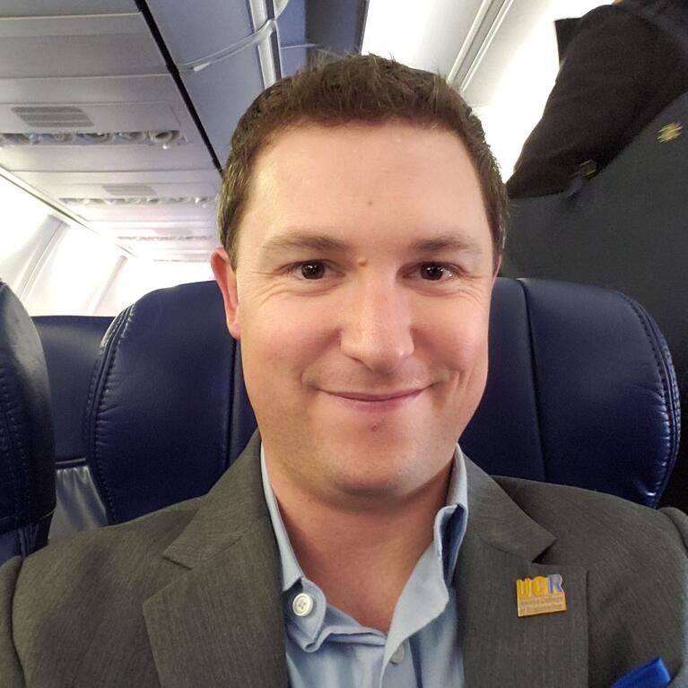 Elliot Emmer, Senior Associate Director of Development, BCOE, gives a warm grin while he sits on an airplane. He is wearing a gray sports jacket with a UCR lapel pin.
