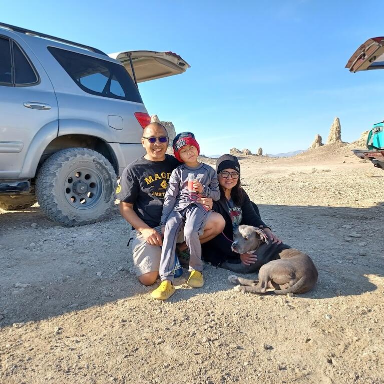 Maite Zabala-Alday poses with her husband, son, and dog at the Mengel Pass in Death Valley, CA. They are sitting on the desert sand and are flanked by two SUVs. 