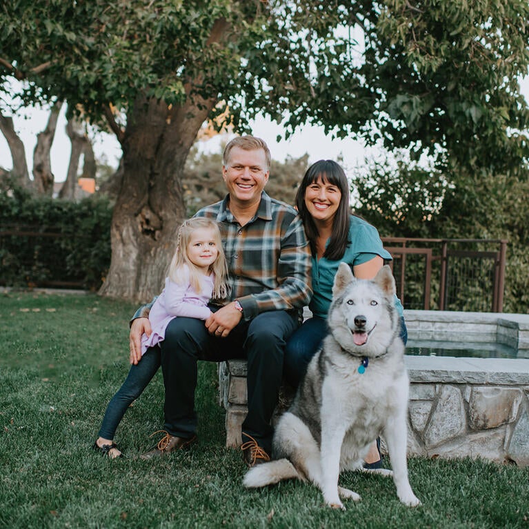 Tracy Telliard poses for a family picture with her husband, daughter and dog. They are seated on a stone bench with green grass on the ground and large trees in the background.