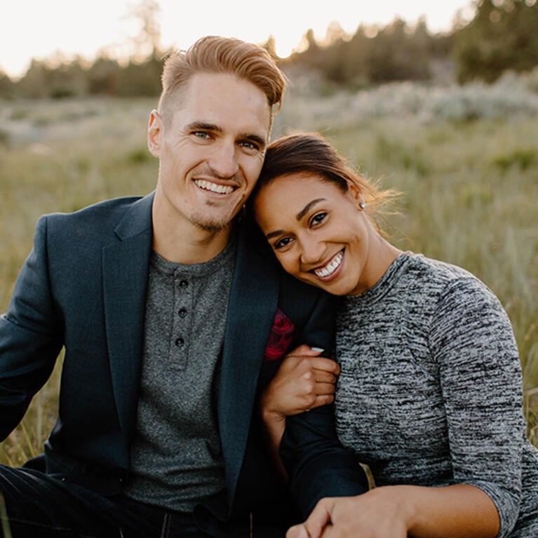 Austin Quick, left, poses in a seated position with his wife, Olivia, in a meadow at sunset. He is wearing a green sports jacket and gray henley T-shirt, while his wife wears a heather gray dress. Austin is the Senior Director of Principal Giving in Development.  