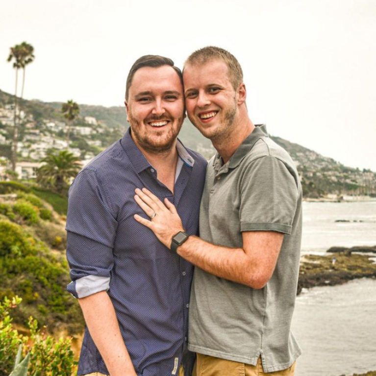 Brock Cavett, left, poses with his fiancé shortly after proposing on the cliffs of Laguna Beach, California. Brock is the Director, Programming and Career Development with Alumni Engagement. Brock is wearing a dark button-down shirt, while his fiancé wears a light polo shirt and has his hands on Brock's chest. The coast and shoreline of Laguna Beach is behind them.