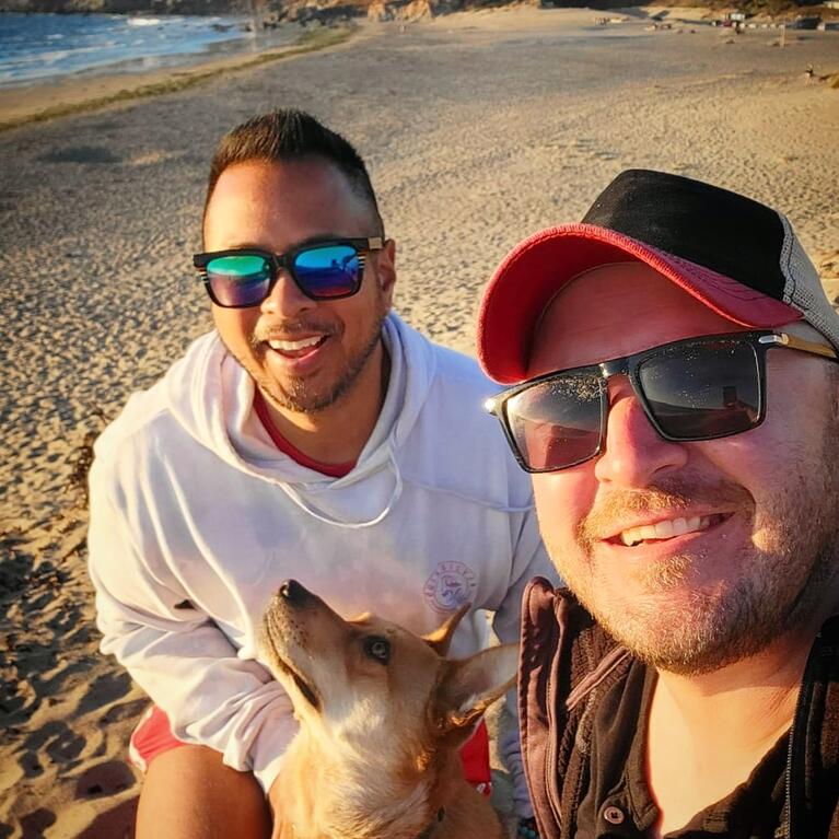 Enrollment marketing manager, Melbert Sebayan, left, poses with his husband and dog. Melbert is wearing reflective sunglasses and a blue hoodie.
