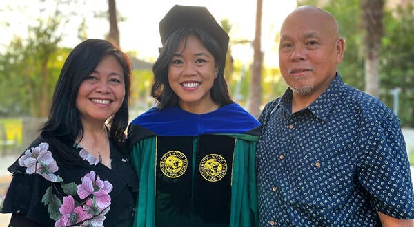Advancement Ally, Christina Magalona, poses in her commencement regalia and is flanked by her parents on either side. There are palm trees in the background.