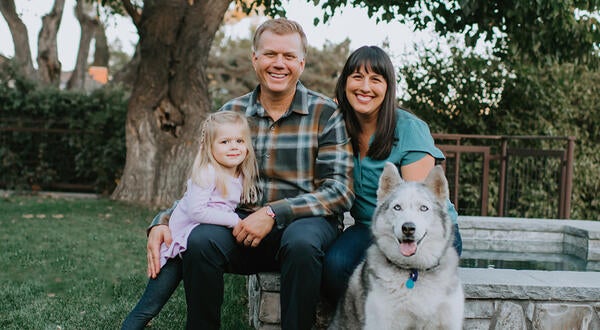 Tracy Telliard poses for a family picture with her husband, daughter and dog. They are seated on a stone bench with green grass on the ground and large trees in the background.