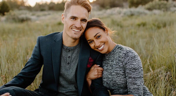 Austin Quick, left, poses in a seated position with his wife, Olivia, in a meadow at sunset. He is wearing a green sports jacket and gray henley T-shirt, while his wife wears a heather gray dress. Austin is the Senior Director of Principal Giving in Development.  