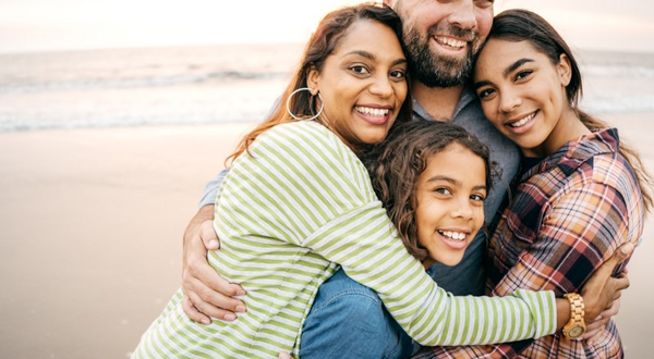 Dalyn Montgomery, Director of Alumni Marketing Strategy & Analytics, poses on the beach with his wife and children in a big group hug.