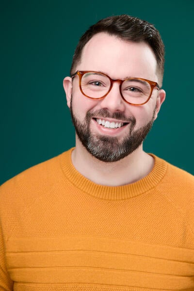 Joshua Carter smiles while wearing an orange sweater and brown glasses while standing agains a hunter green background