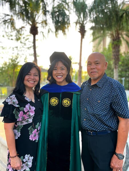 Advancement Ally, Christina Magalona, poses in her commencement regalia and is flanked by her parents on either side. There are palm trees in the background.