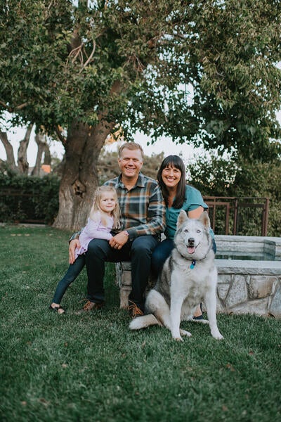 Tracy Telliard poses for a family picture with her husband, daughter and dog. They are seated on a stone bench with green grass on the ground and large trees in the background.