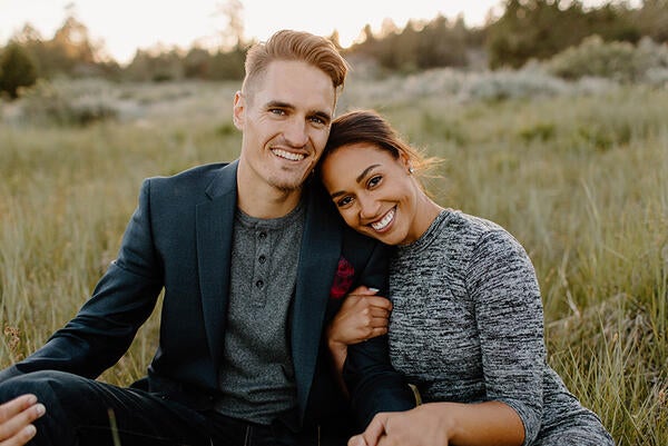 Austin Quick, left, poses in a seated position with his wife, Olivia, in a meadow at sunset. He is wearing a green sports jacket and gray henley T-shirt, while his wife wears a heather gray dress. Austin is the Senior Director of Principal Giving in Development.  