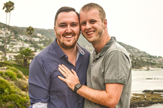 Brock Cavett, left, poses with his fiancé shortly after proposing on the cliffs of Laguna Beach, California. Brock is the Director, Programming and Career Development with Alumni Engagement. Brock is wearing a dark button-down shirt, while his fiancé wears a light polo shirt and has his hands on Brock's chest. The coast and shoreline of Laguna Beach is behind them.