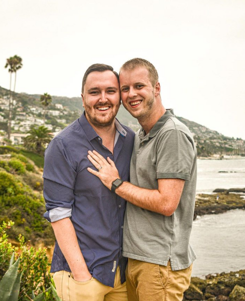 Brock Cavett, left, poses with his fiancé shortly after proposing on the cliffs of Laguna Beach, California. Brock is the Director, Programming and Career Development with Alumni Engagement. Brock is wearing a dark button-down shirt, while his fiancé wears a light polo shirt and has his hands on Brock's chest. The coast and shoreline of Laguna Beach is behind them.