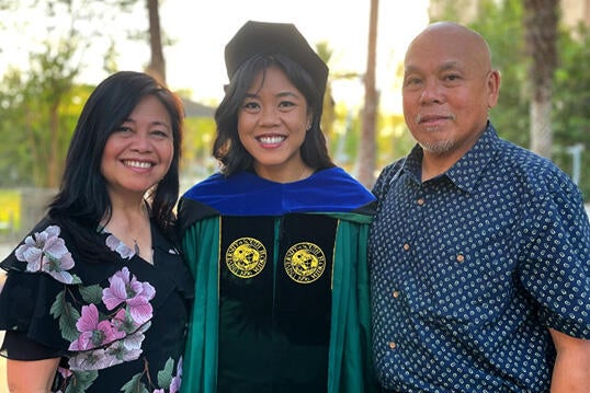 Advancement Ally, Christina Magalona, poses in her commencement regalia and is flanked by her parents on either side. There are palm trees in the background.