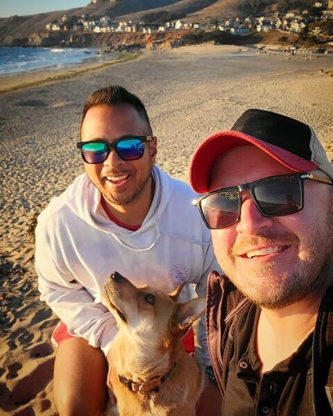 Enrollment marketing manager, Melbert Sebayan, left, poses with his husband and dog. Melbert is wearing reflective sunglasses and a blue hoodie.