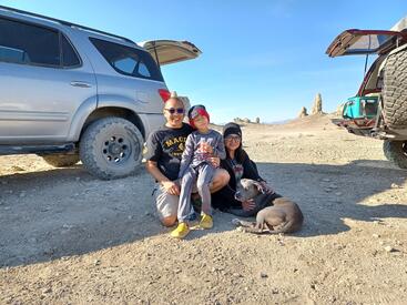 Maite Zabala-Alday poses with her husband, son, and dog at the Mengel Pass in Death Valley, CA. They are sitting on the desert sand and are flanked by two SUVs. 