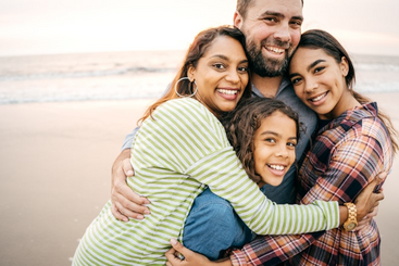 Dalyn Montgomery, Director of Alumni Marketing Strategy & Analytics, poses on the beach with his wife and children in a big group hug.